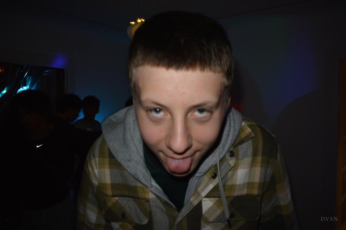 A young man leans toward the lens under direct flash, sticking out his tongue, a dim party behind him.