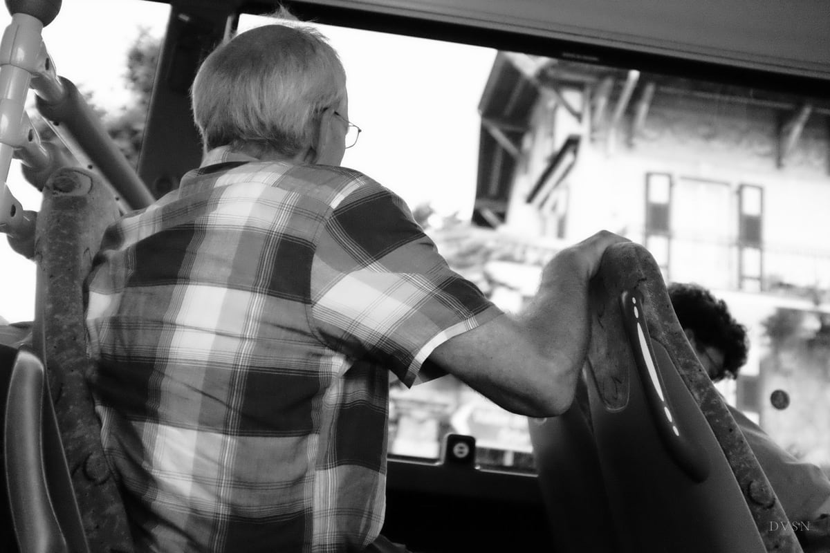 An older man in a checked shirt, seen from behind, holding a seat on a bus.