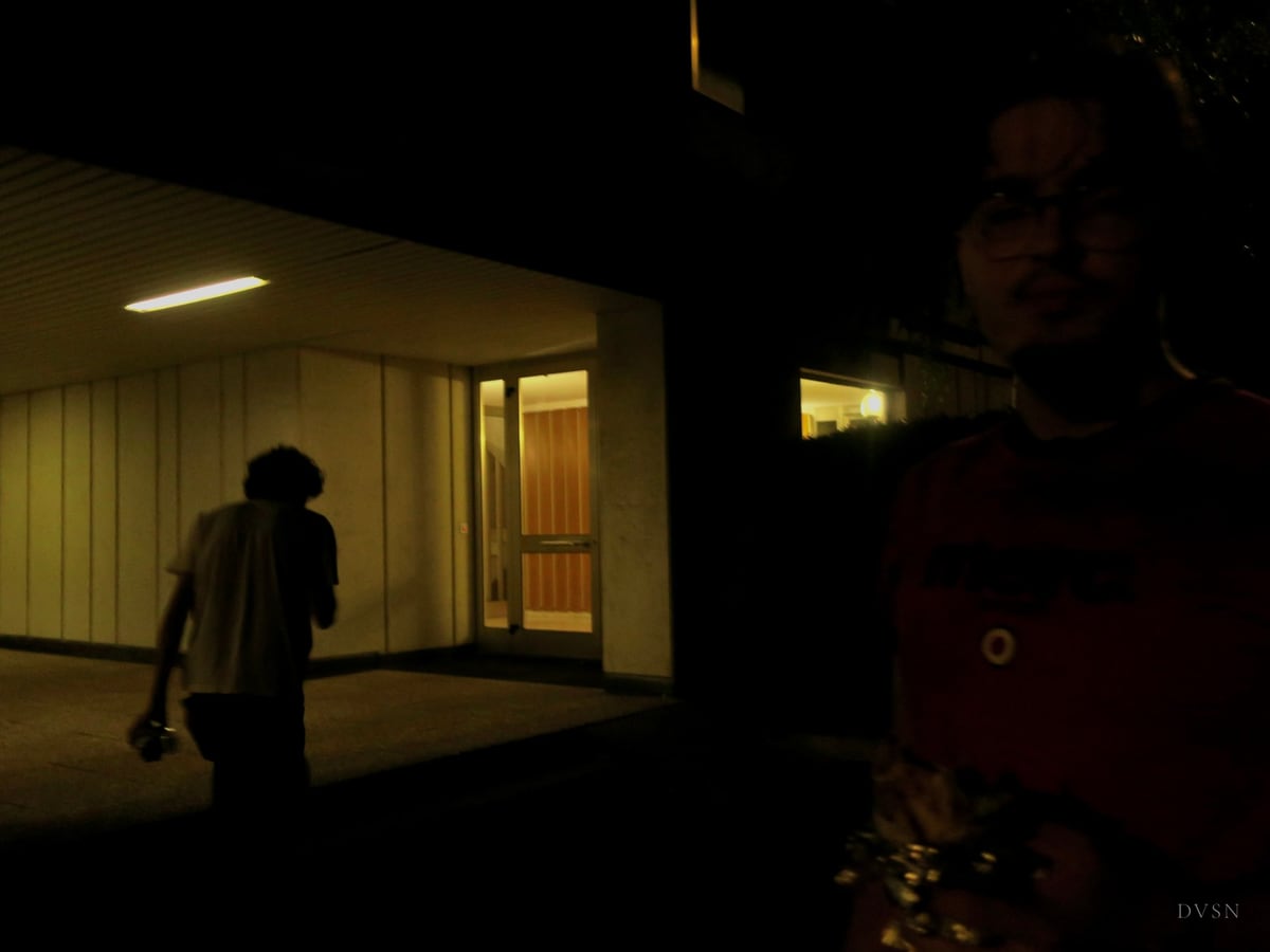 A silhouette walks toward a lit entrance at night while a half-lit face watches from the right edge.