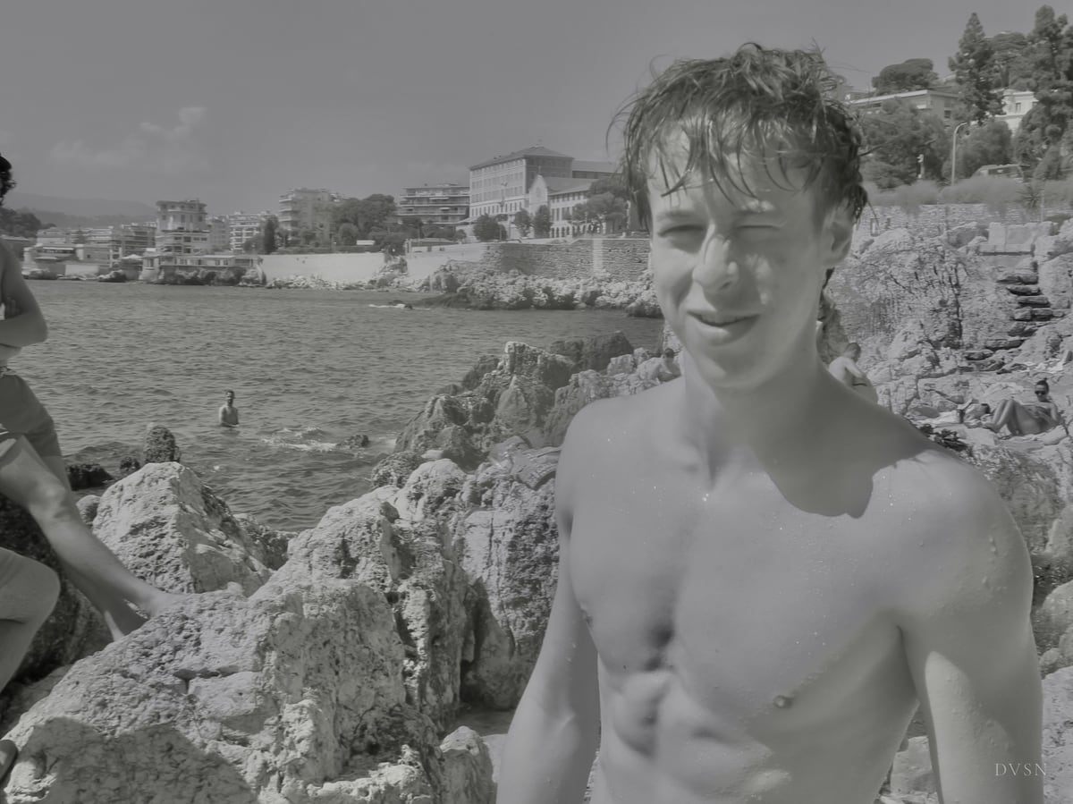 A young man, wet-haired and shirtless, smiling on seaside rocks.