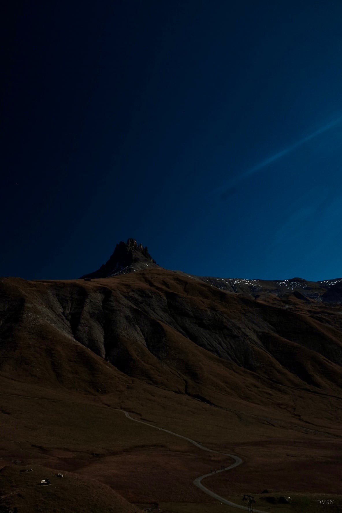 A rocky peak against a deep blue sky above dark slopes, a winding path and tiny walkers below.