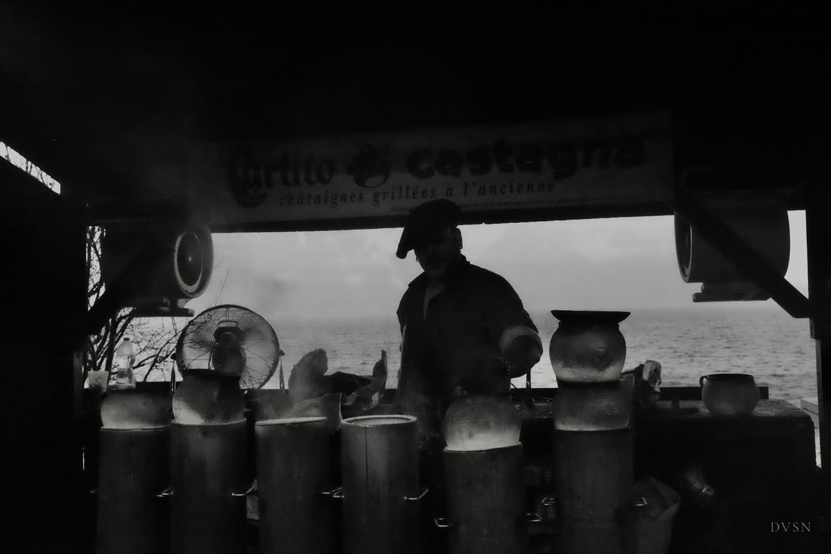 A chestnut vendor in silhouette behind his roasting stand, steam rising, pale water and sky beyond.
