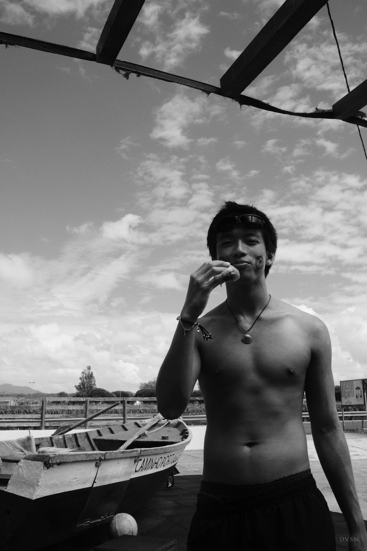 A shirtless young man eating under the beams of a pergola, a small boat beside him and a wide sky above.