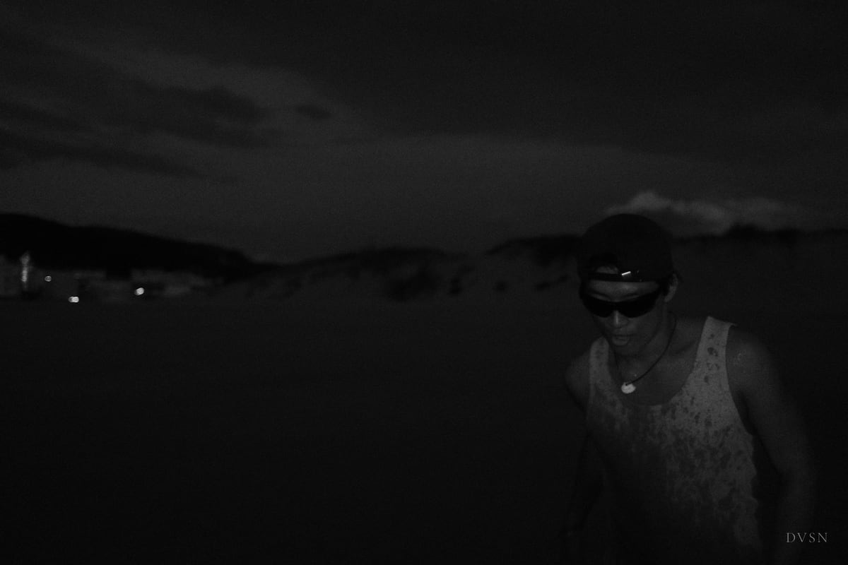 A man in a cap and sunglasses emerges from near-total darkness by the shore at night.