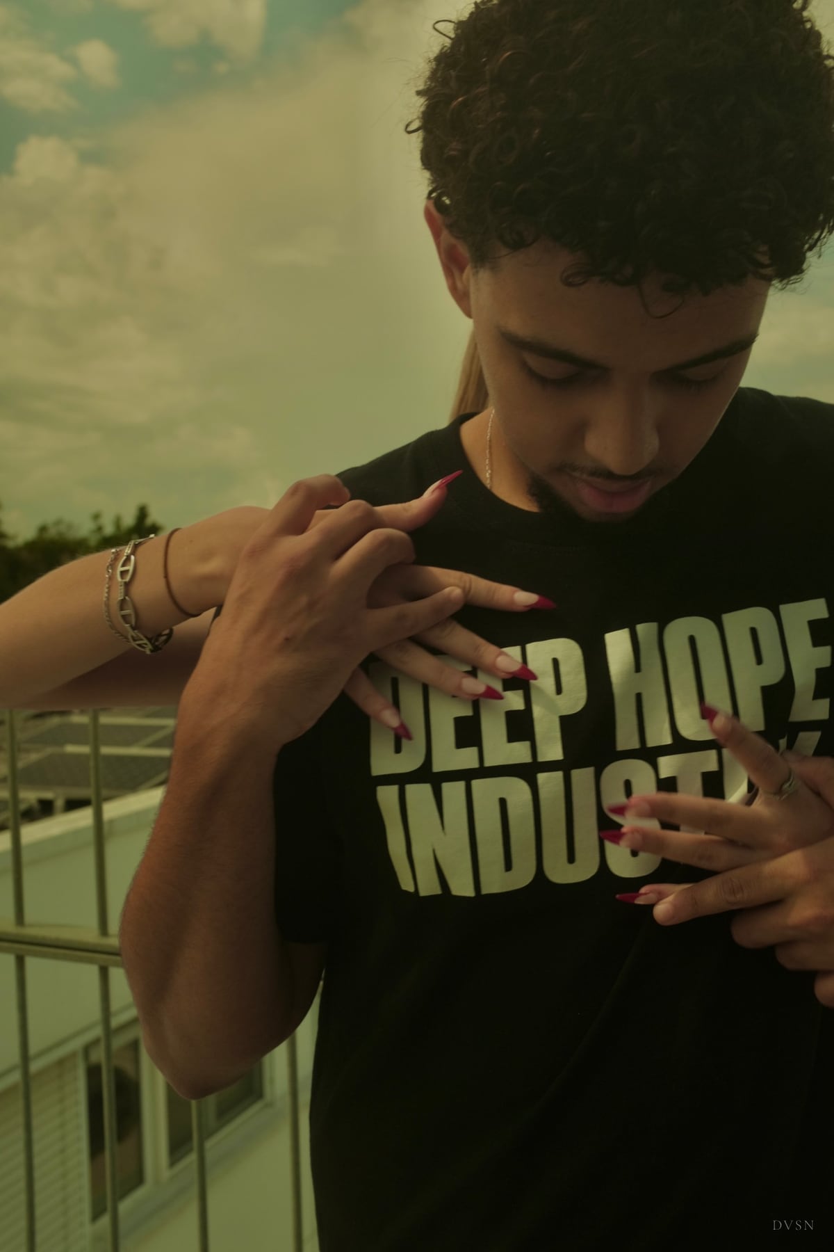 A young man looks down as hands with long red nails reach across his black printed T-shirt from behind.