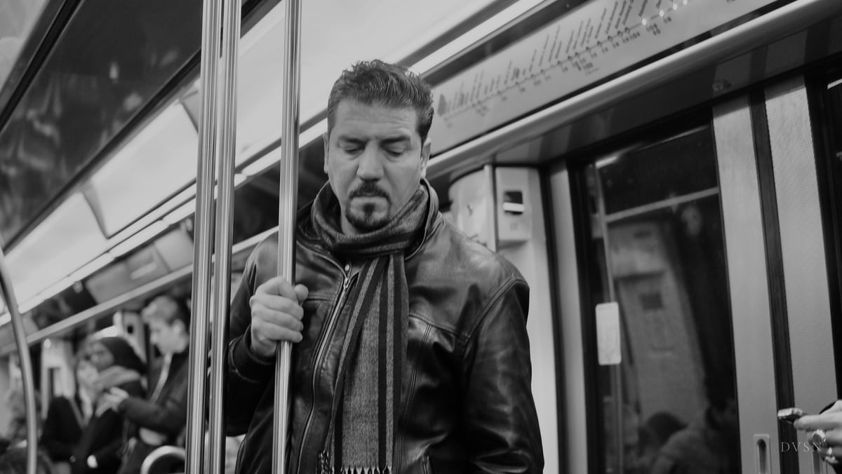 A man in a leather jacket and scarf holds a pole in a metro carriage, eyes lowered.