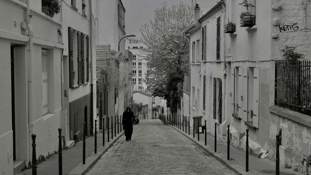 A lone figure in a long dark coat walks down a cobbled street lined with bollards.