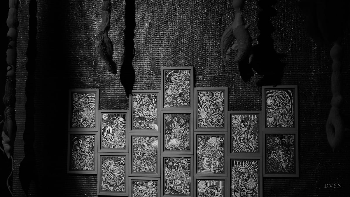 A wall of framed black-and-white drawings beneath hanging textile forms, in dim light.