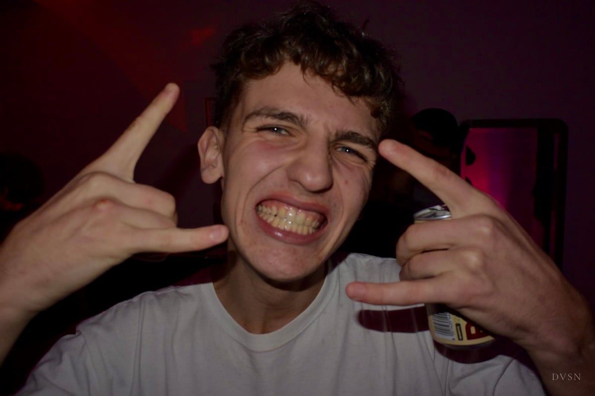 A young man grins at the camera under flash, hands raised in a playful gesture, a can in one hand.