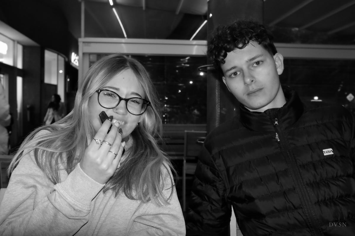 Two friends at a café terrace at night; she laughs behind her hand, he stares calmly into the lens.