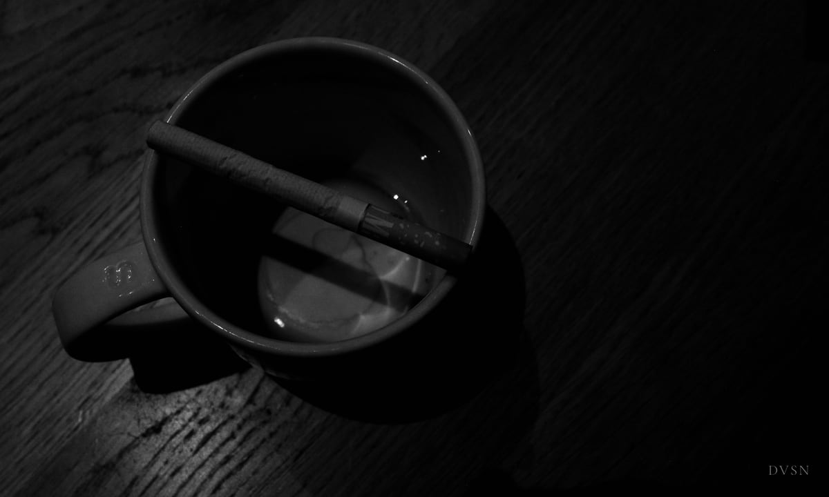 A cigarette resting across an empty mug on a dark wooden table, seen from above.