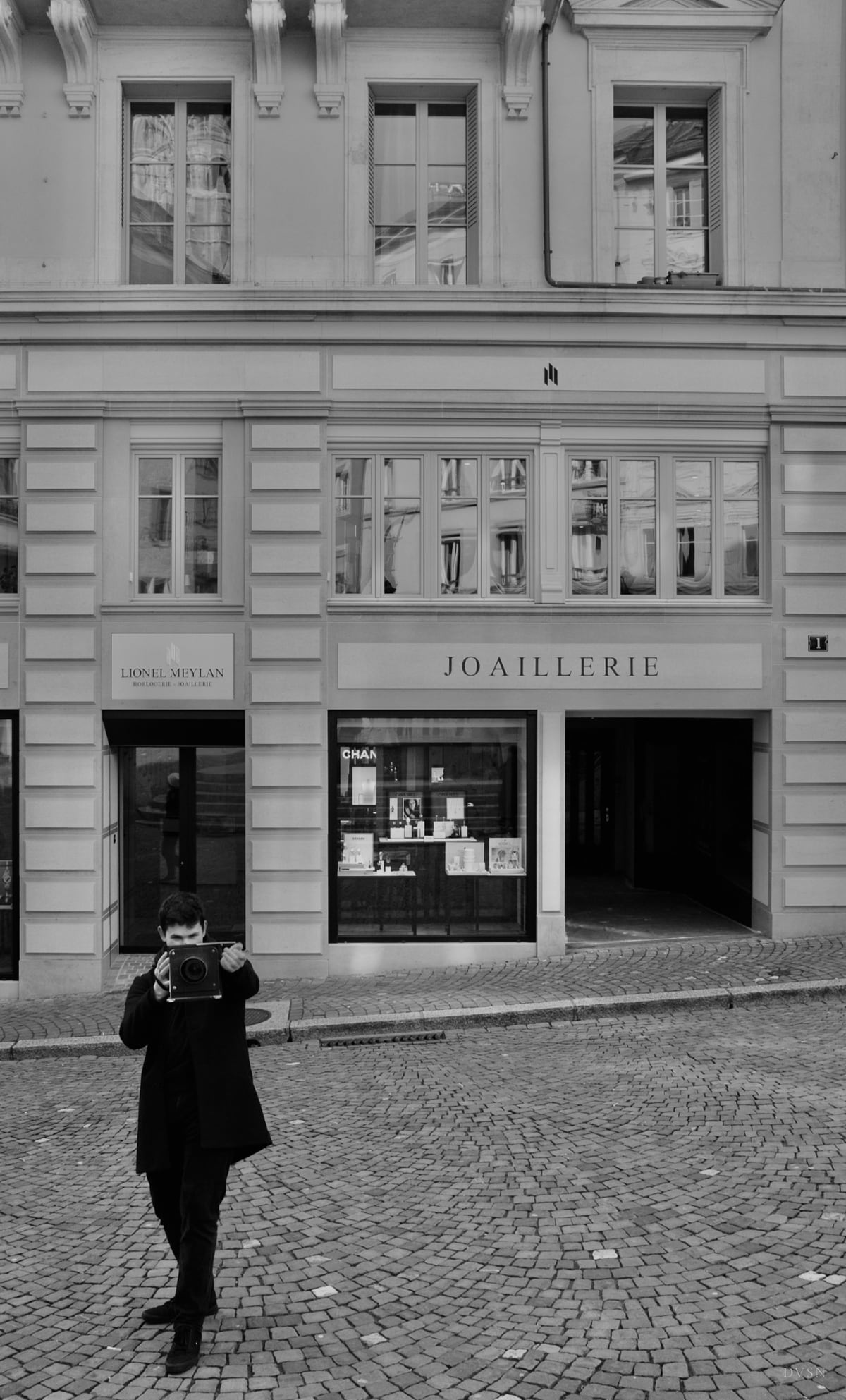 A man in a dark coat points a large-format camera at the viewer, in front of a stone façade with a jewellery shop.